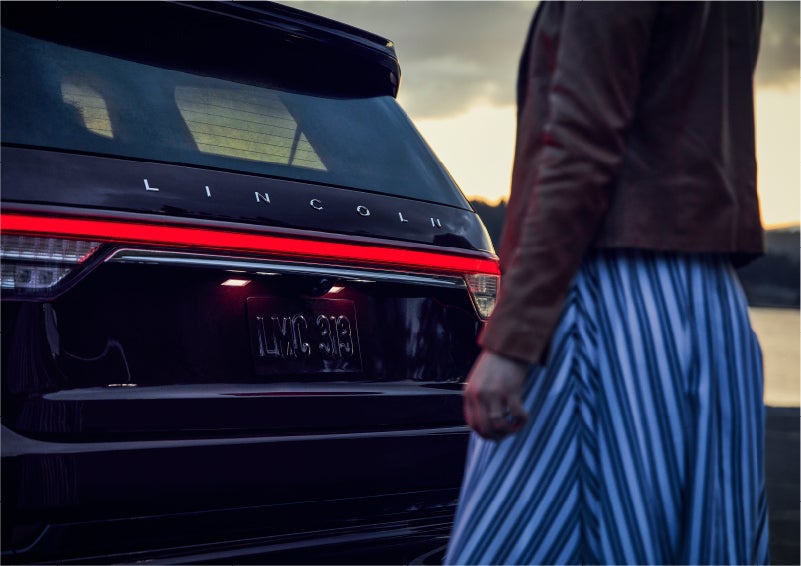A person is shown near the rear of a 2023 Lincoln Aviator® SUV as the Lincoln Embrace illuminates the rear lights | Pierre Lincoln in Lynnwood WA